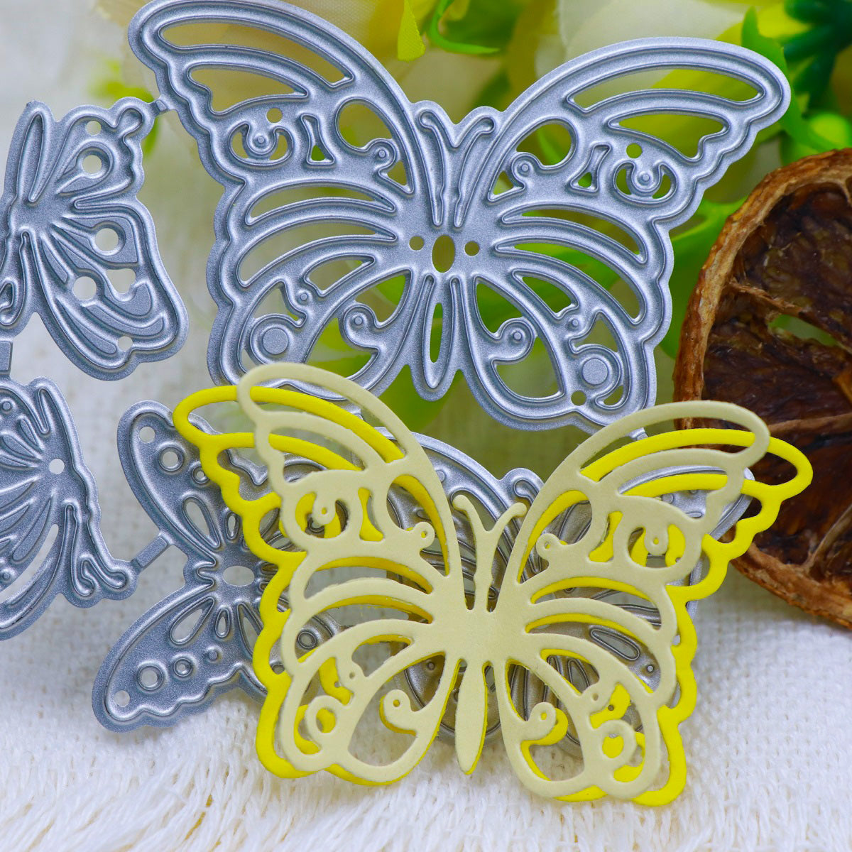 Metal and yellow paper butterfly-shaped cutouts, crafted with the Printable Heaven Fluttering Butterflies Cutting Die (5PCS) (A214), are beautifully arranged on a white textured surface, accented by green and white flowers and a dried brown slice in the background—ideal inspiration for your next cardmaking supplies collection.
