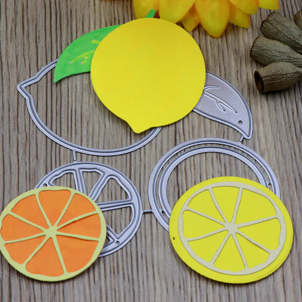 Paper cutouts of vibrant citrus fruit slices, made using the Printable Heaven Lemon Citrus Fruit Cutting Die (4 PCS) (L098) and other coordinating dies, are artfully displayed on a wooden surface alongside a yellow flower and dried seed pods—perfect inspiration for your next paper crafting project.