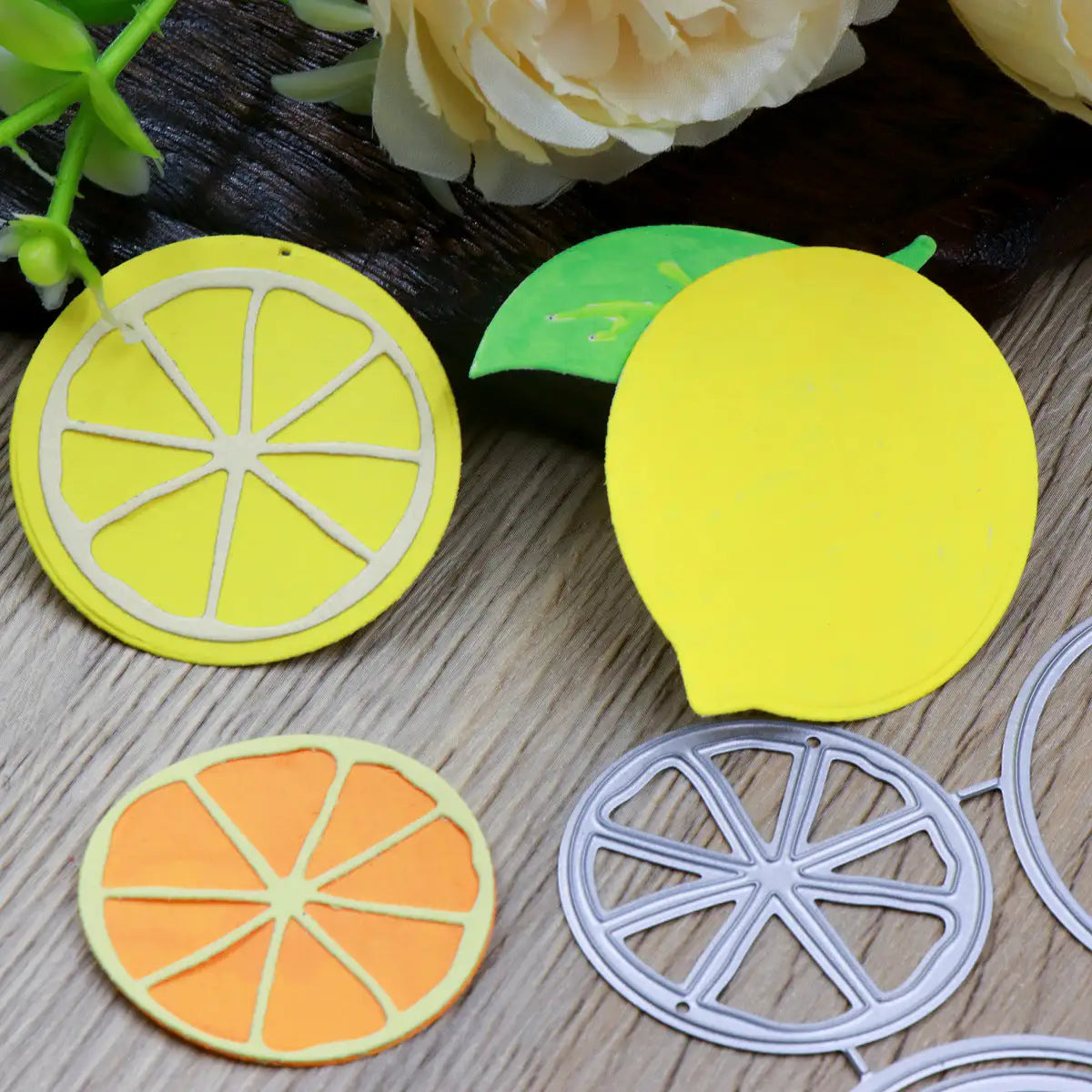 Vibrant paper cutouts of lemon and orange slices, made using the Printable Heaven Lemon Citrus Fruit Cutting Die (4 PCS) (L098), are artfully arranged on a wooden surface beside white flowers—creating a lively scene ideal for paper crafting projects.