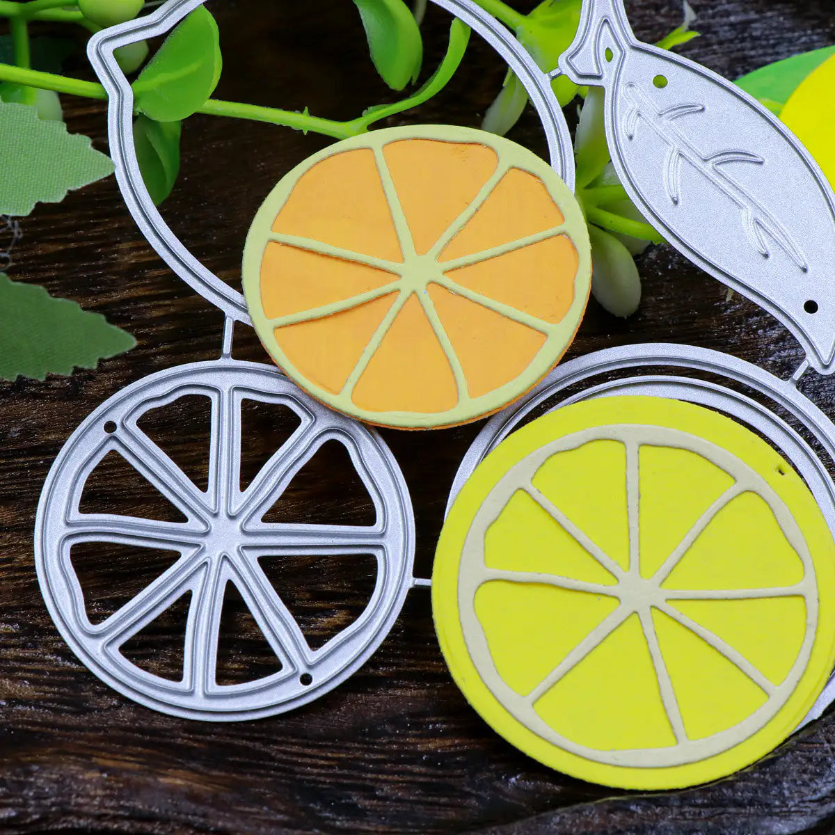Printable Heaven’s Lemon Citrus Fruit Cutting Die (4 PCS) (L098) set features intricately designed dies to create citrus slices and leaves—including a detailed lemon slice—which are beautifully showcased alongside finished orange and yellow paper die-cut fruits. These vibrant embellishments, ideal for paper crafting projects, are artfully arranged on a dark, textured wooden surface with green artificial foliage in the background.