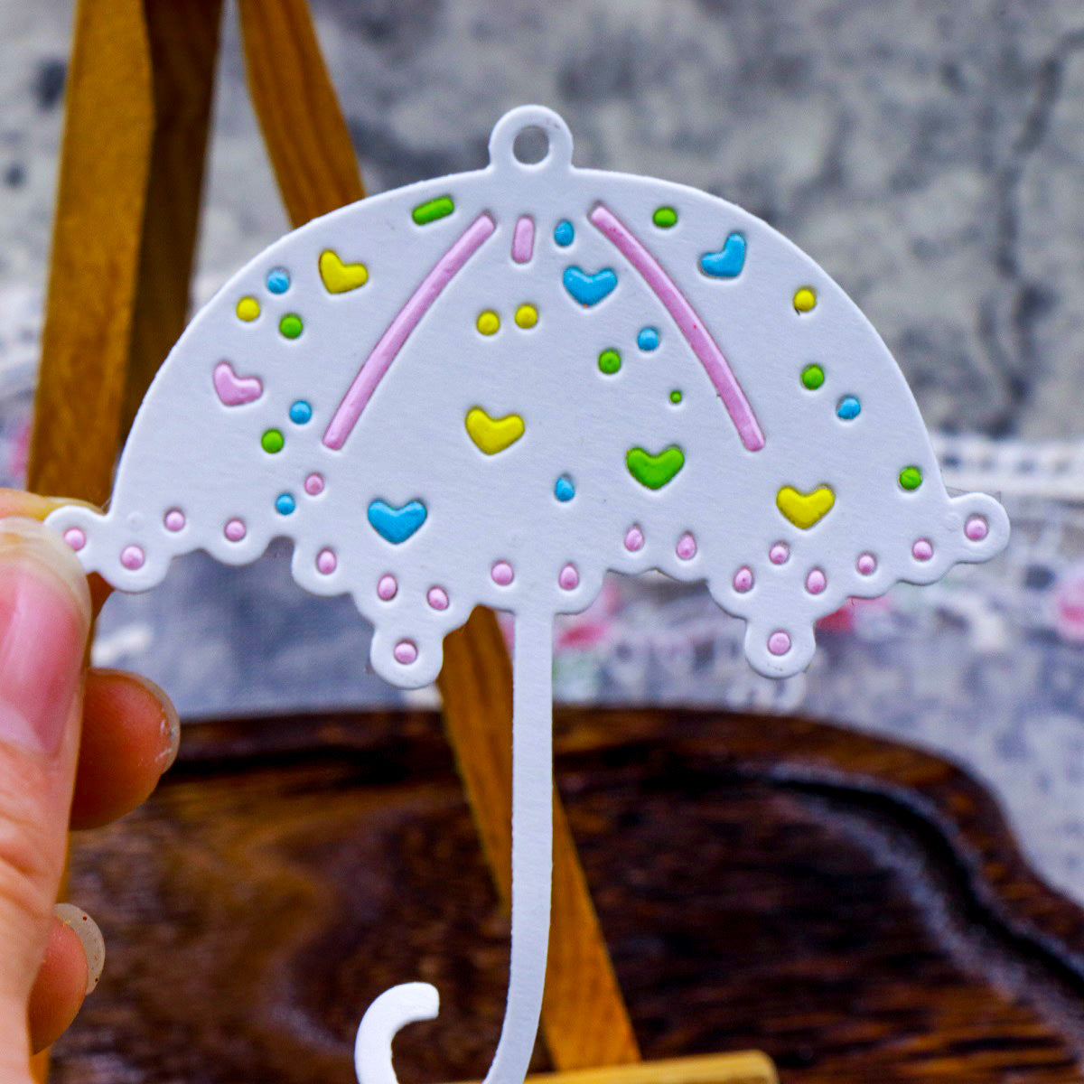 A hand holds a white decorative paper umbrella embellished with colorful heart and dot patterns, set against a softly blurred wooden and fabric backdrop—an inspiring piece for paper craft lovers and card makers, evoking the Parasol Umbrella Cutting Die (1 PC) (D073) by Printable Heaven.