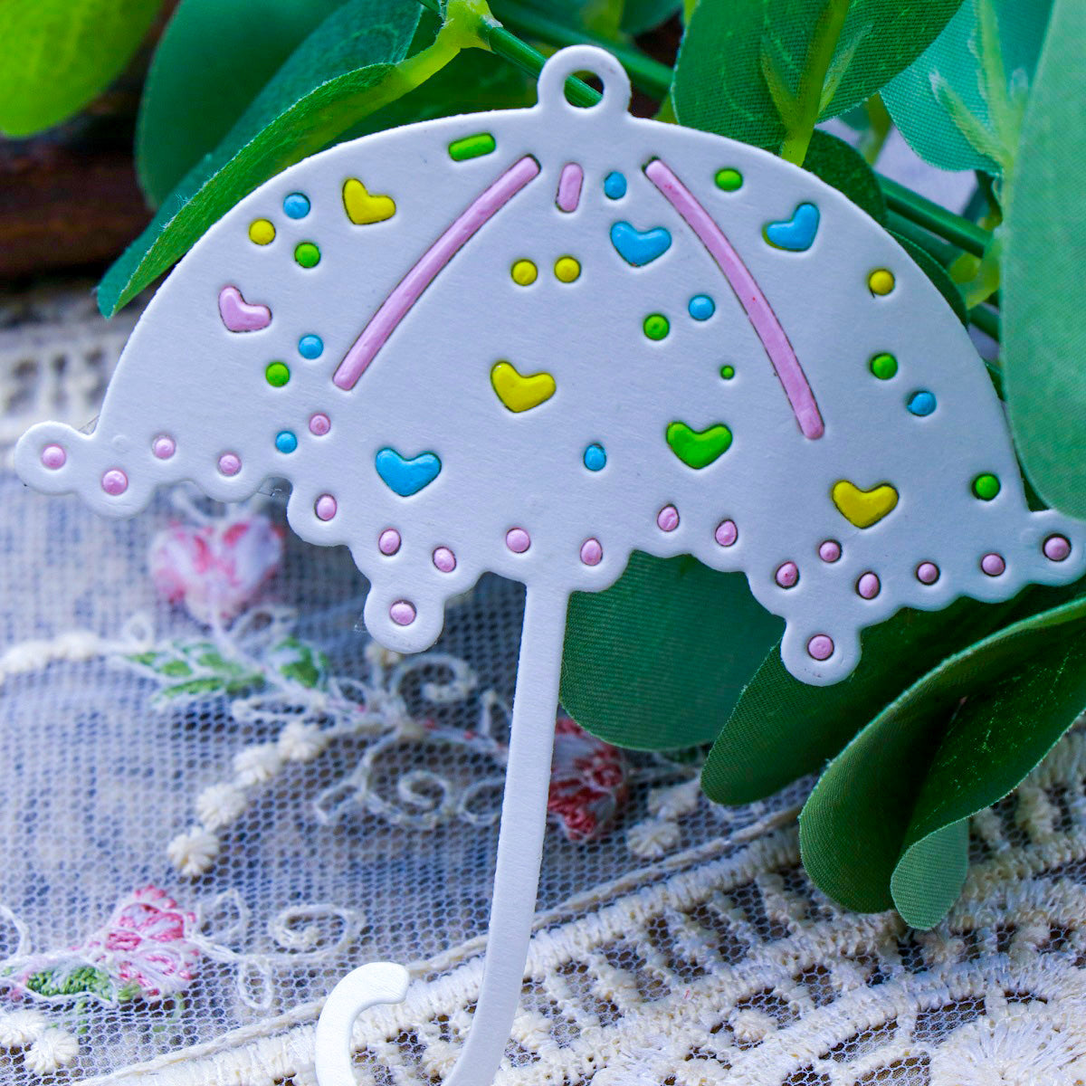 A stylish Parasol Umbrella Cutting Die (1 PC) (D073) by Printable Heaven features a decorative metal design with colorful heart-shaped accents, shown resting on lace fabric and surrounded by green leaves and floral embroidery—ideal inspiration for card makers or anyone looking for unique paper craft ideas using this versatile cutting die.