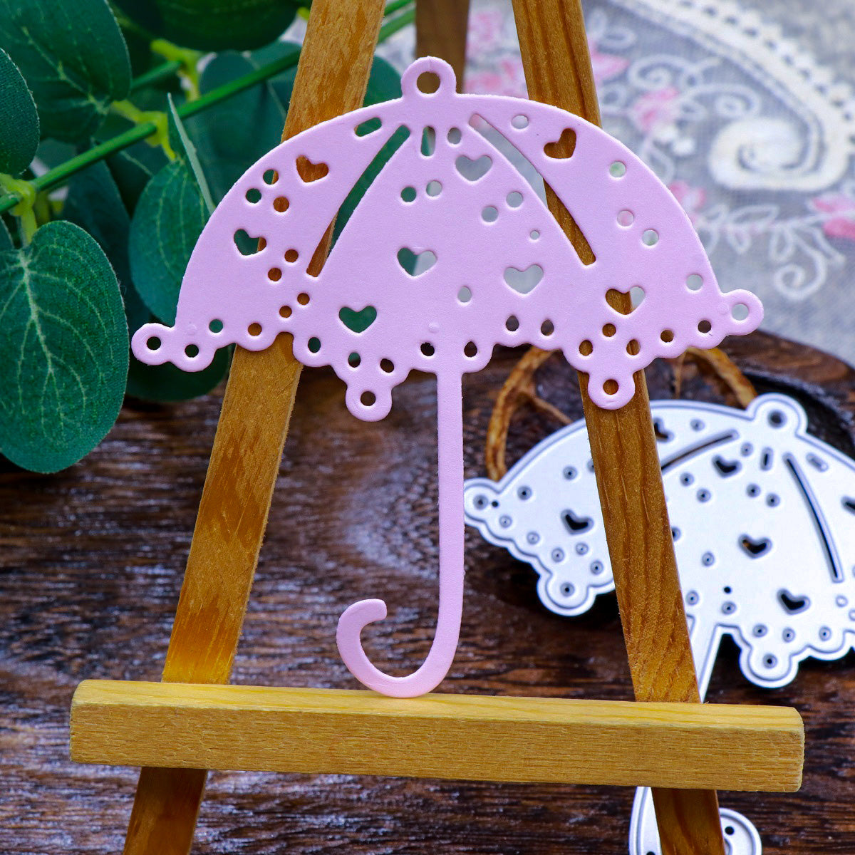 A pink, heart-patterned umbrella decoration—ideal for card making and other papercrafts—crafted with the Printable Heaven Parasol Umbrella Cutting Die (1 PC) (D073), is displayed on a small wooden easel, accompanied by a leafy plant and a matching silver Parasol Umbrella Cutting Die piece in the background.