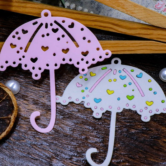 Two decorative umbrella cutouts created using the Printable Heaven Parasol Umbrella Cutting Die (1 PC) (D073)—one in pink and another in white adorned with colored hearts—are displayed on a wooden surface among a dried orange slice, pearls, and patterned fabric. Ideal for paper crafting and card making projects.