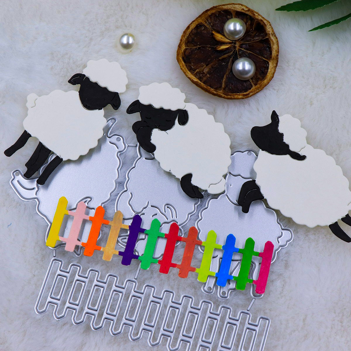Four sheep and lamb-shaped cutting dies from the Printable Heaven “Trio of Little Lambs with Picket Fence Border (4pcs) (A063)” set are arranged on a furry background, with a vibrant mini picket fence and a dried orange slice adorned with two pearls nearby—ideal inspiration for Easter cards or charming farmyard-themed crafts.