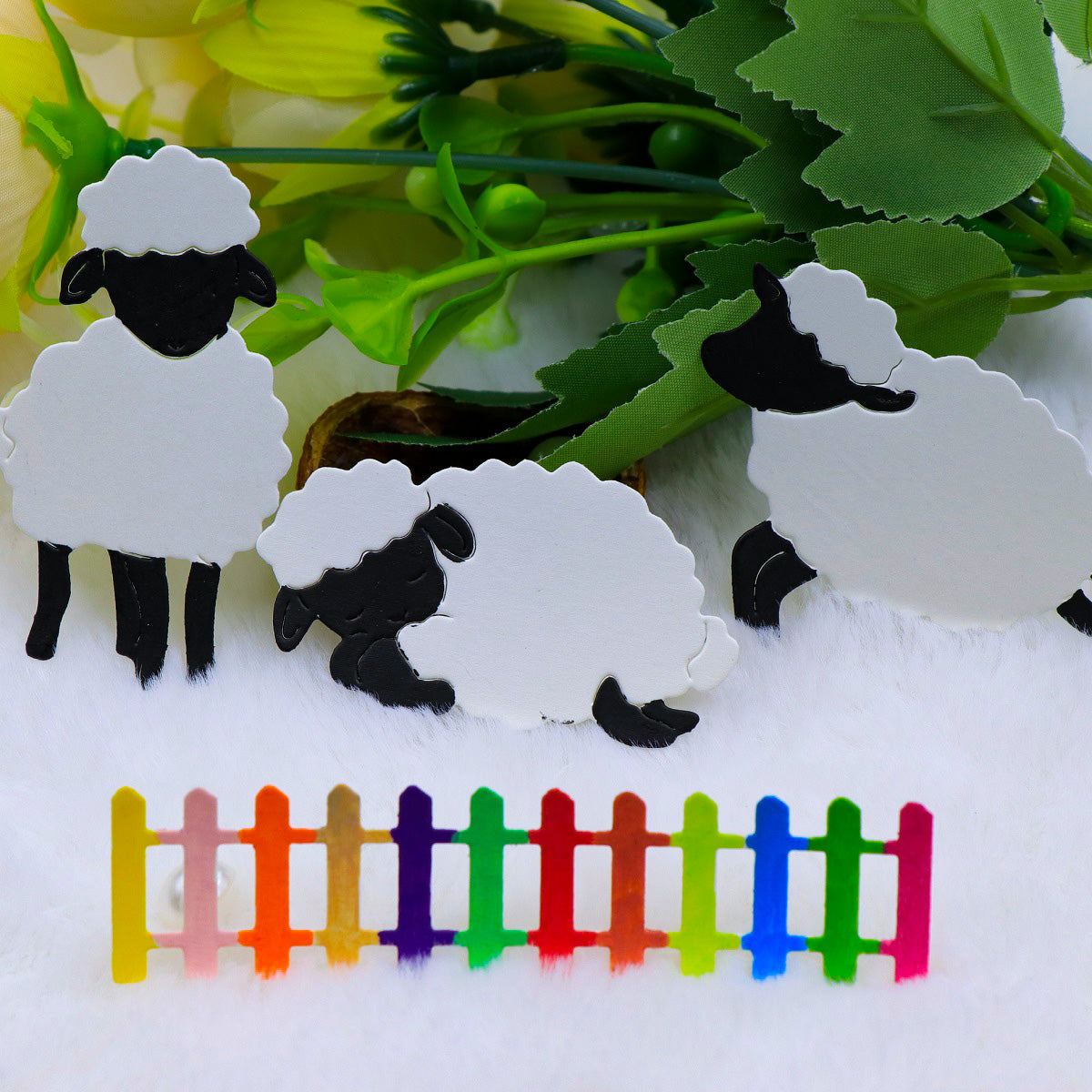 Three adorable lamb figures from the Printable Heaven “Trio of Little Lambs with Picket Fence Border (4pcs) (A063)” set—featuring black-faced, white-bodied sheep arranged on a white surface with artificial green leaves and yellow flowers—are displayed alongside a small, colorful picket fence. This charming arrangement is perfect inspiration for Easter card making or creative farmyard crafts.