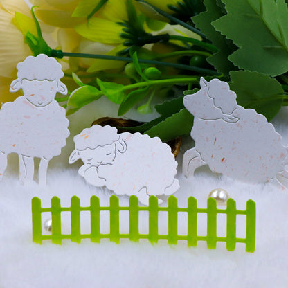 Three white paper cutouts from Printable Heaven’s “Trio of Little Lambs with Picket Fence Border (4pcs) (A063)” set stand on white fabric, with greenery and yellow flowers in the background. Perfect for farmyard crafts or Easter card making, a small green picket fence from the set and two pearls are arranged in front of the sheep.