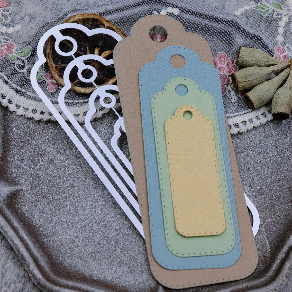 Four layered, rectangular paper tags in beige, green, blue, and yellow with rounded tops—perfectly crafted using the Printable Heaven Stitched Edge Tags Die Set (4 PCS) (SP068)—are stacked on a lace and velvet surface near dried fruit and flowers. The distinctive stitched edge outlines created by the die set are also visible underneath.