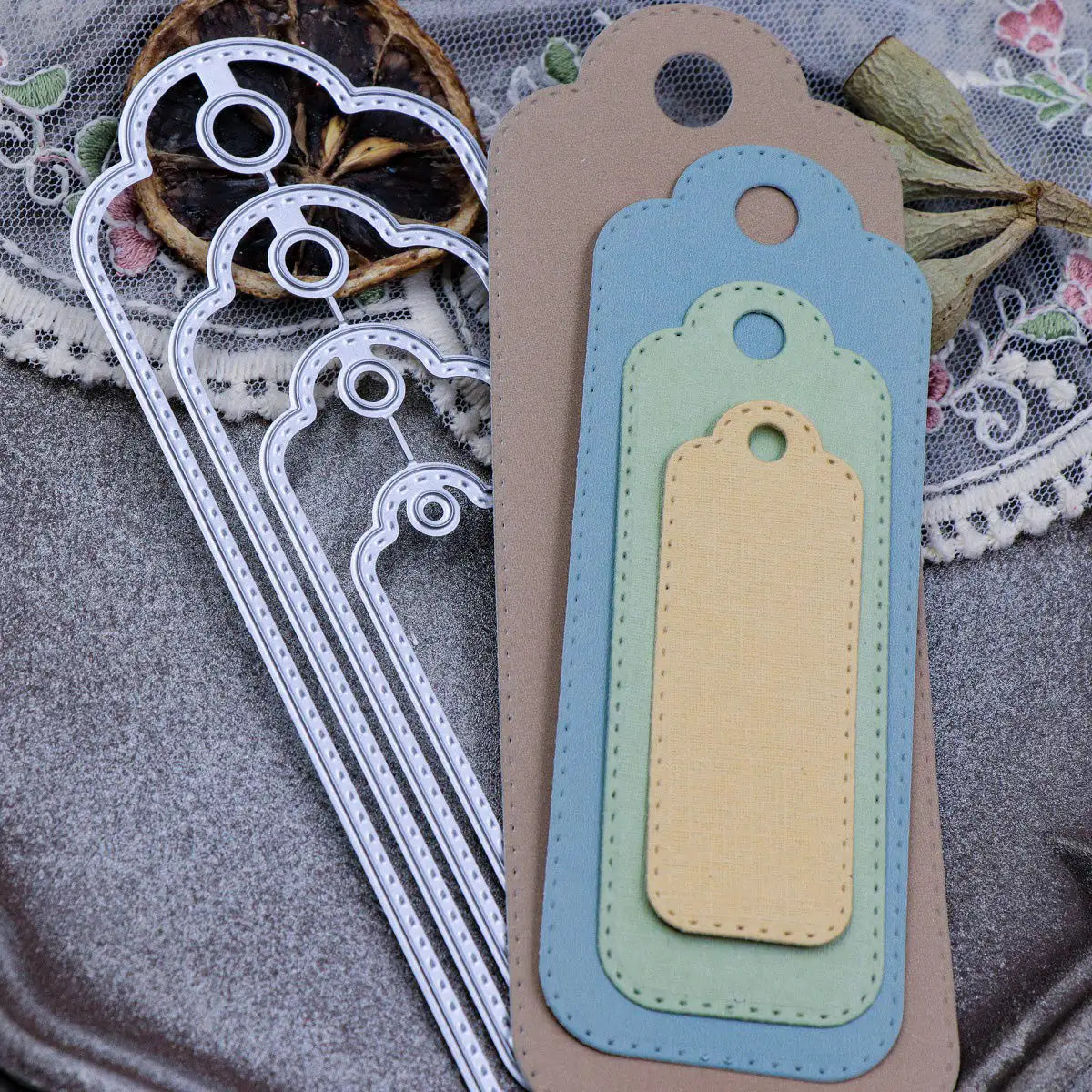 The Printable Heaven Stitched Edge Tags Die Set (4 PCS) (SP068) is beautifully showcased with four layered paper tags in brown, blue, and yellow hues, displayed alongside nesting tag dies. Arranged on a lace tablecloth with dried flowers, this charming setup is perfect for anyone passionate about paper crafting.