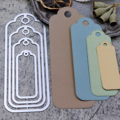 Four layered metal tag dies are shown next to four cut paper tags in brown, blue, green, and beige, all with rounded tops and holes. Perfect for paper crafting projects, these nesting tag dies feature a delicate stitched edge. Dried flowers and slices are arranged in the background on a textured surface.