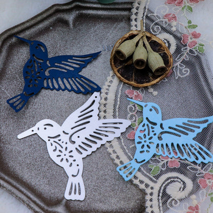 Three paper hummingbirds in navy, white, and light blue—crafted with the Printable Heaven Hummingbird Metal Cutting Die (1 PC) (A116) using die-cutting machines for papercraft projects—are arranged on a silver tray alongside a lace cloth, dried seed pods, and floral embroidery.