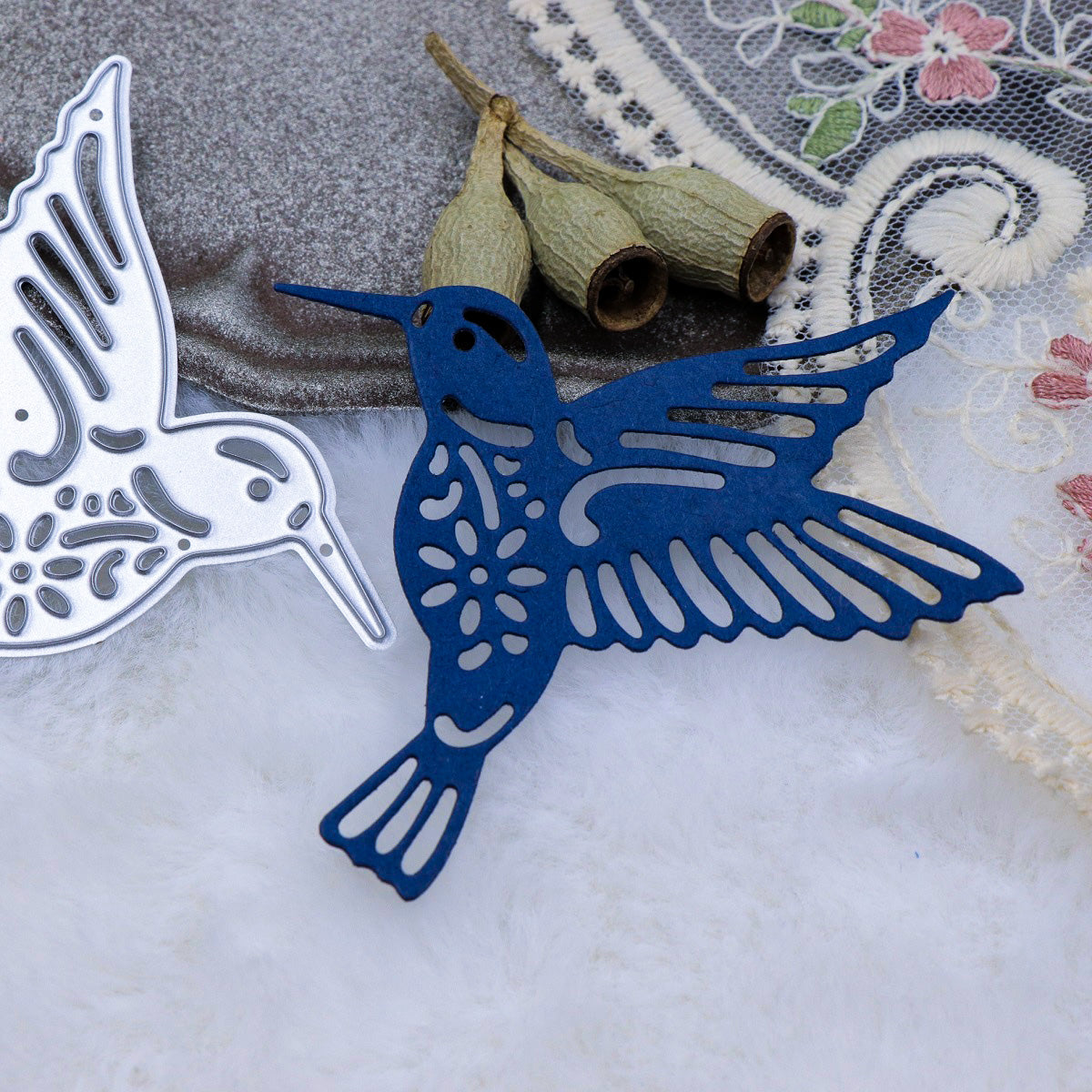 Two decorative hummingbird cutouts, made with the Printable Heaven Hummingbird Metal Cutting Die (1 PC) (A116) and ideal for papercraft projects, rest in silver and dark blue on a white furry surface near dried seed pods and floral-embroidered fabric.