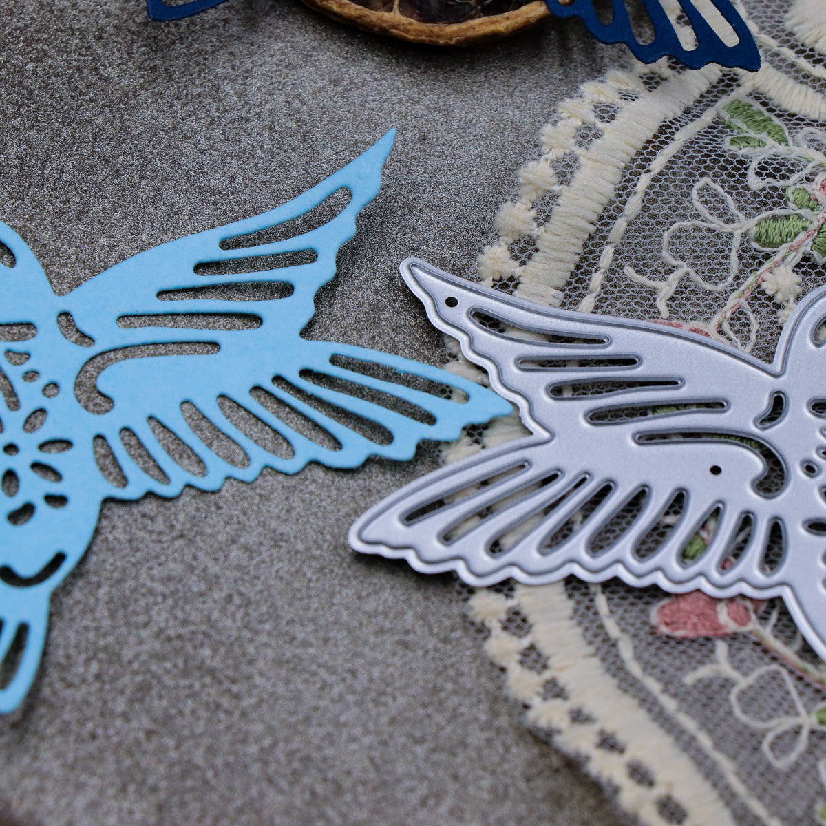 Close-up of two intricately cut bird-shaped decorations, crafted using the Printable Heaven Hummingbird Metal Cutting Die (1 PC) (A116), displayed on a textured surface next to lace-trimmed fabric—a perfect inspiration for papercraft projects.