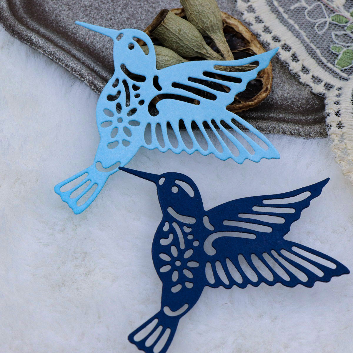 Two intricately cut paper hummingbirds, one light blue and one dark blue, are displayed on a white surface near dried leaves and a piece of embroidered fabric—perfect accents for papercraft projects made using the Hummingbird Metal Cutting Die (1 PC) (A116) by Printable Heaven.