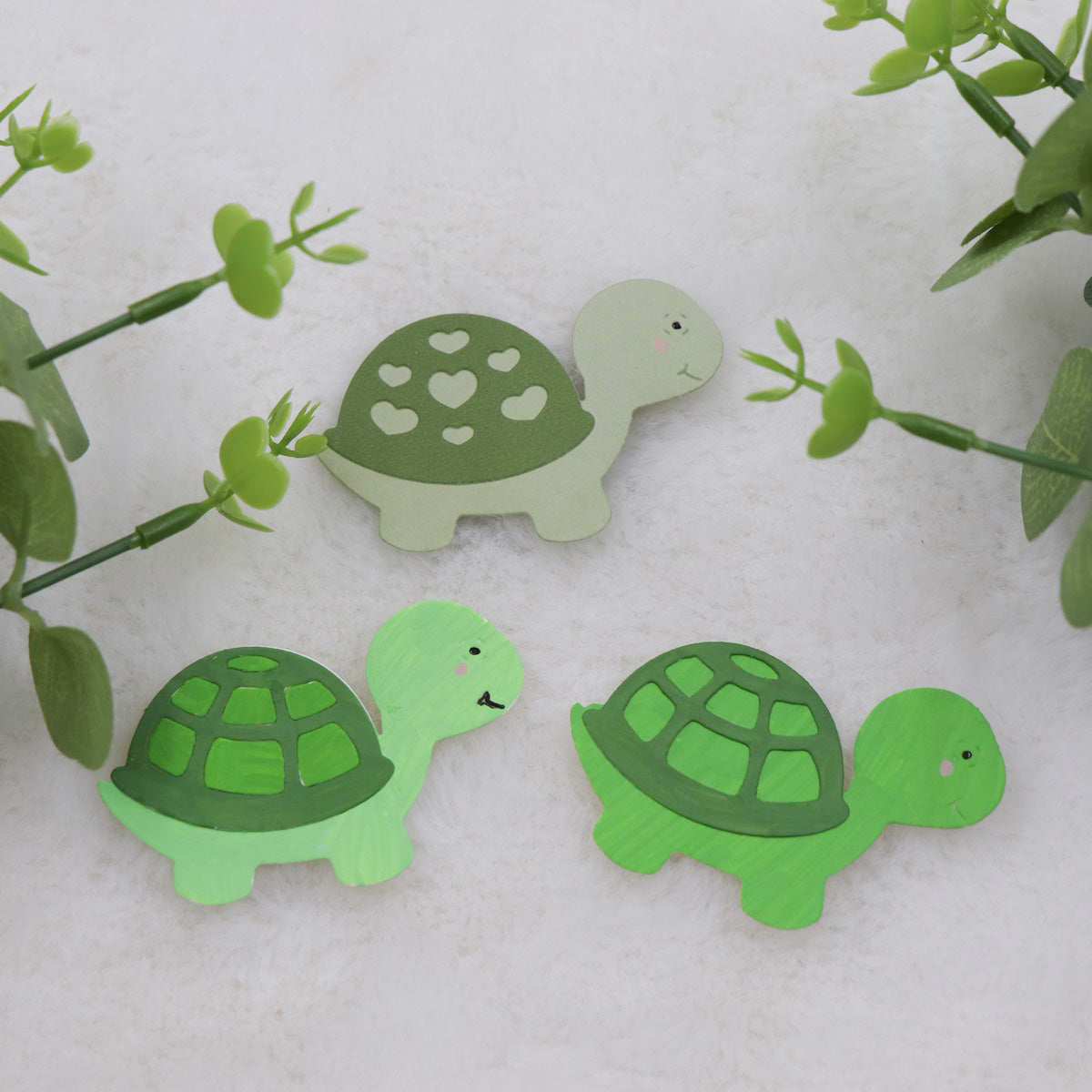 Three cheerful green cartoon turtle cutouts rest on a white surface, surrounded by lush leafy plants. Created with the Printable Heaven Turtle/Tortoise Cutting Die (3 PCS) (A203), these charming die-cuts feature uniquely decorated shells—one with heart shapes and the others with classic patterns—making them ideal for card making or scrapbooking projects.