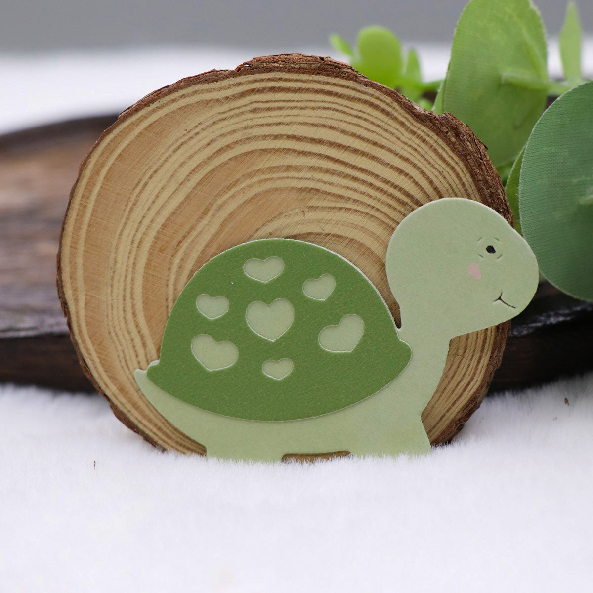 A wooden slice stands upright with an adorable green turtle cutout—decorated with heart-shaped patterns on its shell—displayed in front. Crafted using the Printable Heaven Turtle/Tortoise Cutting Die (3 PCS) (A203), this piece is ideal for card making or scrapbooking. Green leaves serve as a decorative background.