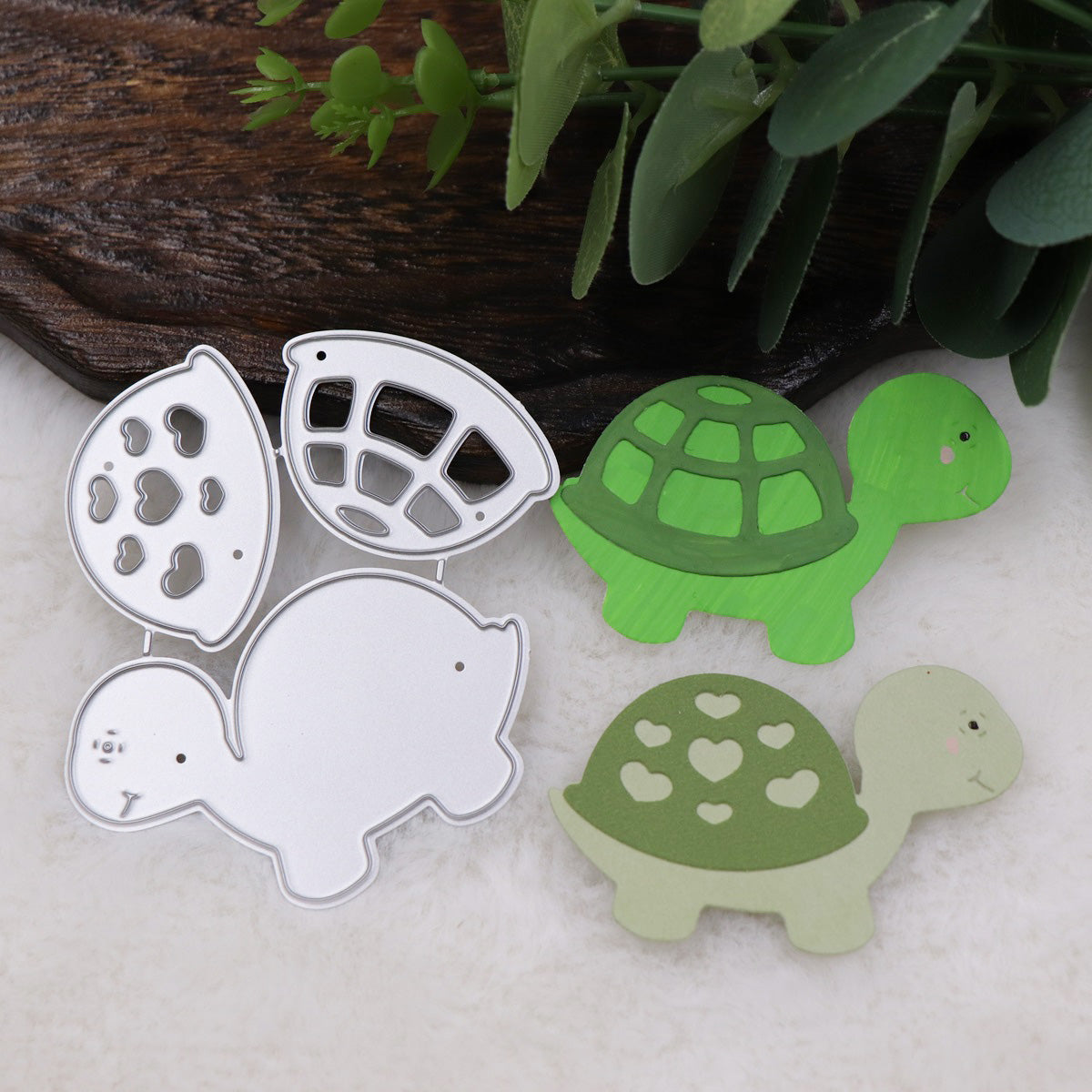 Three turtle-shaped cutouts, created using the Turtle/Tortoise Cutting Die (3 PCS) (A203) from Printable Heaven, are shown on white fabric next to green leaves and a wooden surface. The cutouts—two made of metal and one of paper—display heart and circle patterns on their shells, making them perfect for scrapbooking and card making.
