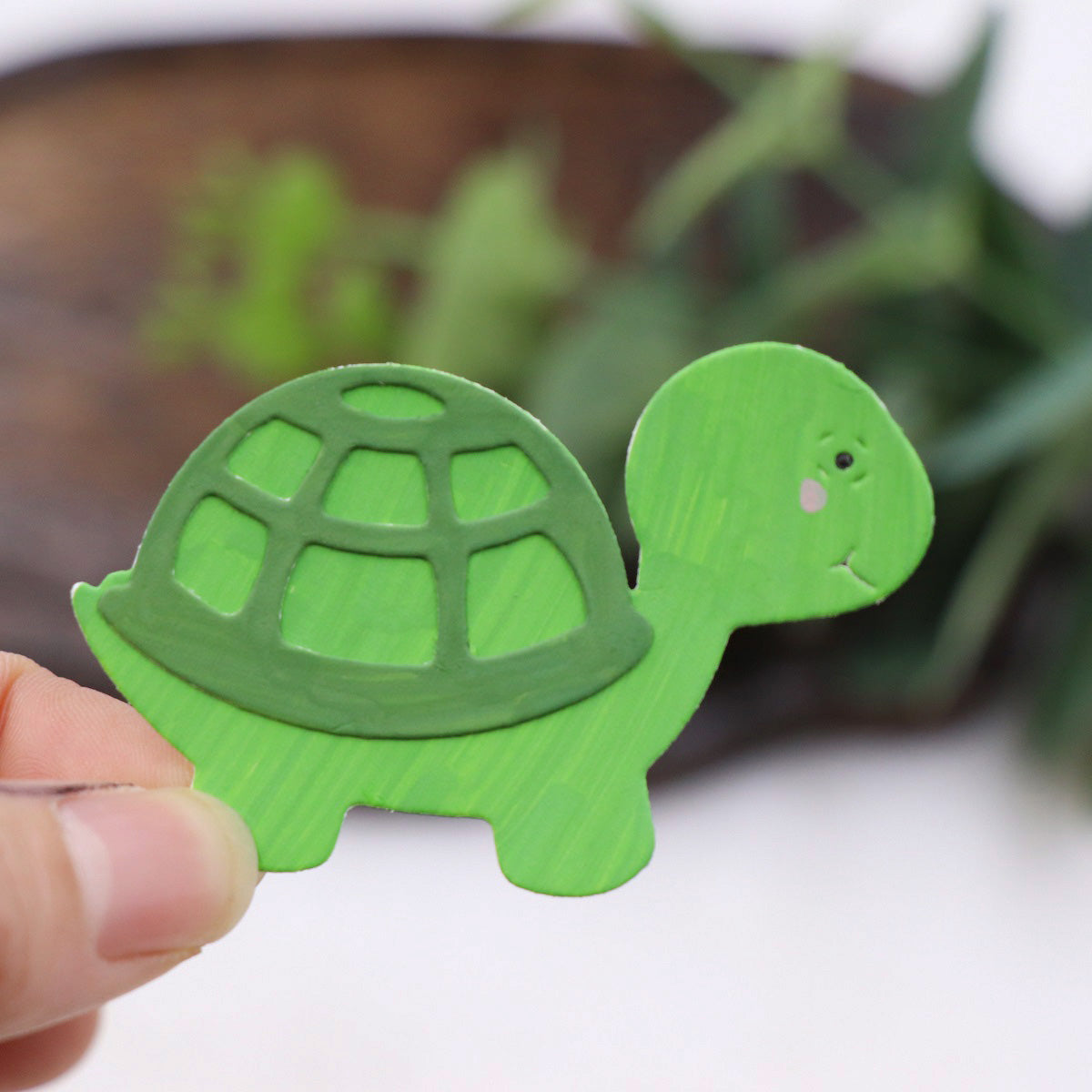 A hand holds a small, green turtle-shaped cutout with a lighter green shell design—perfect for card making or scrapbooking. The blurred background features leafy plants, emphasizing the intricate details created using the Printable Heaven Turtle/Tortoise Cutting Die (3 PCS) (A203).