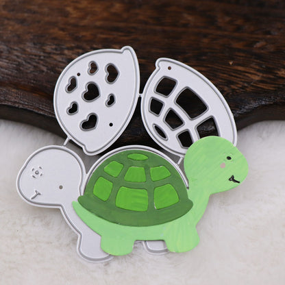 A Printable Heaven Turtle/Tortoise Cutting Die set (3 PCS) (A203) is displayed, featuring a silver metal turtle-shaped die positioned behind a green and light green turtle cutout—both resting on a white, fluffy surface with a piece of dark wood in the background. Perfect for card making or scrapbooking.