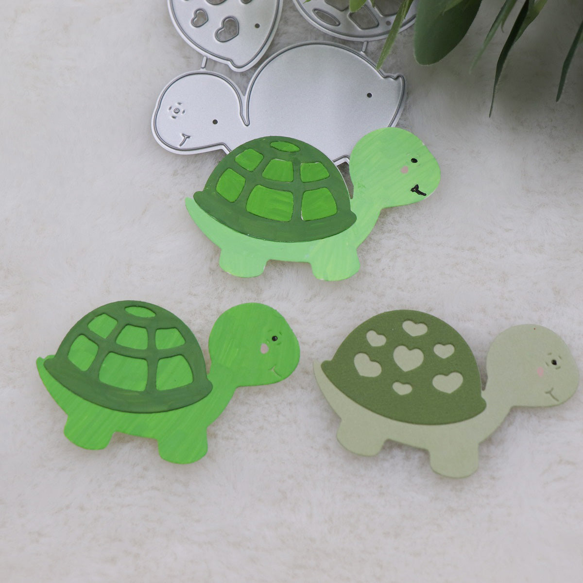 Featuring the Printable Heaven Turtle/Tortoise Cutting Die (3 PCS) (A203), this scene displays three charming turtle-shaped cutouts: two are vibrant green with classic shell patterns, and one is light green adorned with heart shapes on its shell. The metal cutting die is visible in the background along with leafy accents—making these delightful scrapbook embellishments ideal for card making and creative projects.