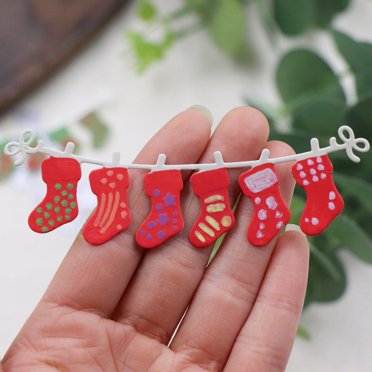 A hand displays the Printable Heaven Christmas Stocking Garland Cutting Die (1 PC) (SP050), featuring a miniature string of six red stockings with unique, colorful patterns—an adorable handmade decoration perfect for Christmas crafting.