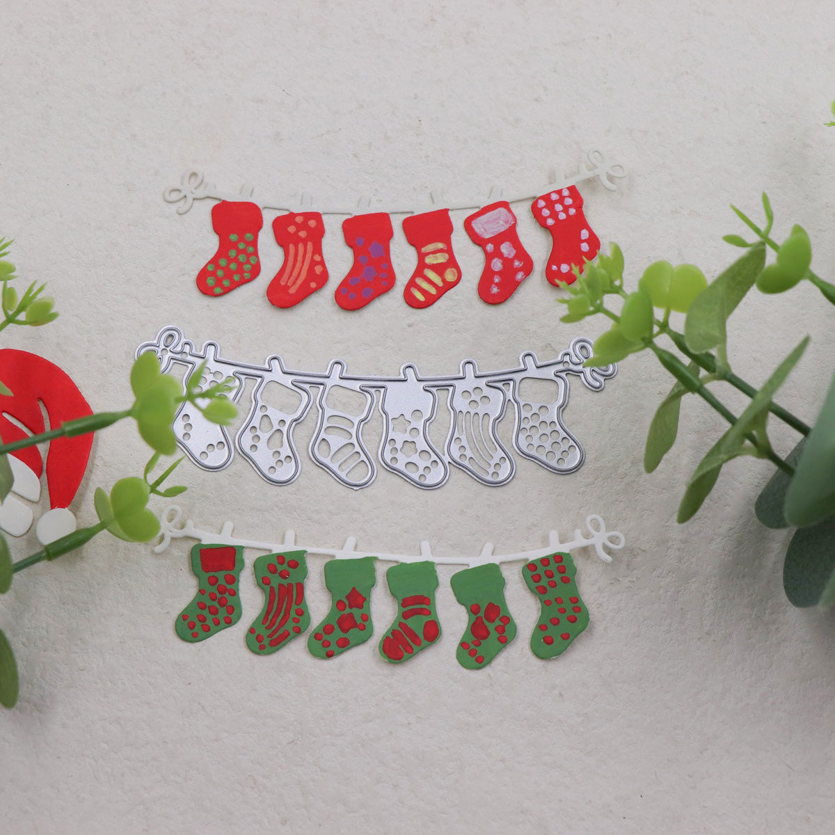 Three garlands made with the Christmas Stocking Garland Cutting Die (1 PC) (SP050) by Printable Heaven—red, silver, and green—hang on a light surface surrounded by green plants. Each garland showcases small, patterned stockings in festive holiday colors and designs, ideal for displaying your handmade decorations or for anyone who enjoys Christmas crafting.