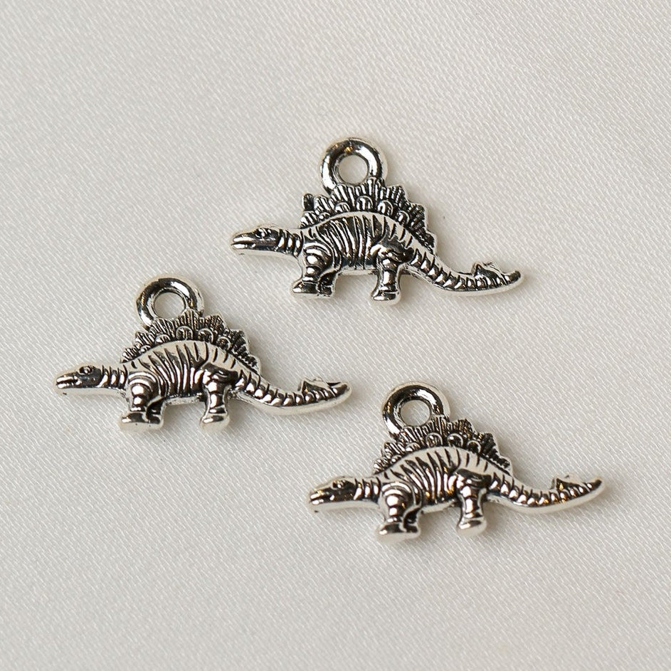 Three silver-toned Stegosaurus Metal Charms (3pcs) (MLB-073) shaped like stegosauruses with loops for hanging.