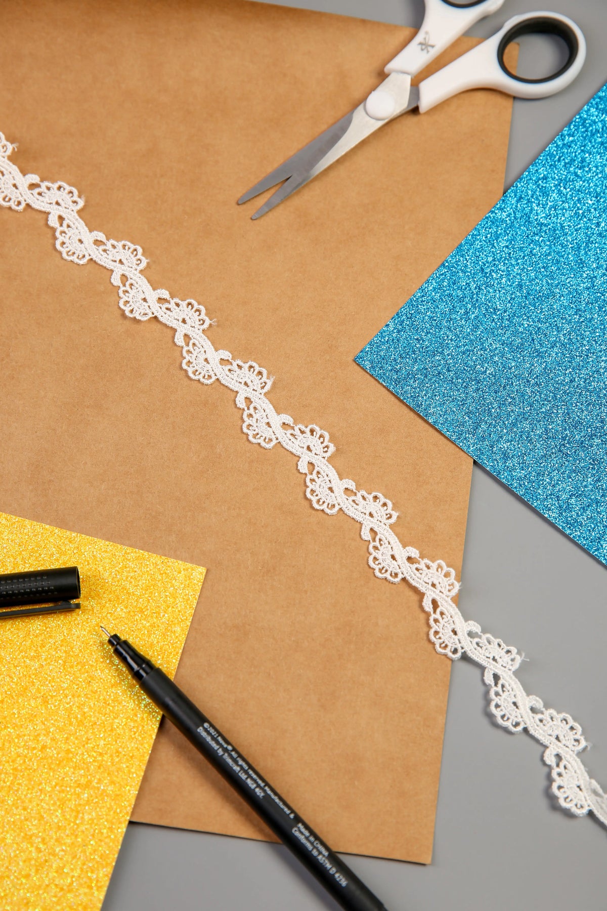 Brown paper, blue and yellow glitter paper, Wavy Floral Lace Edging (RT10458 IVORY), scissors, and two black markers are arranged on a flat surface, suggesting a crafting or DIY project setup.