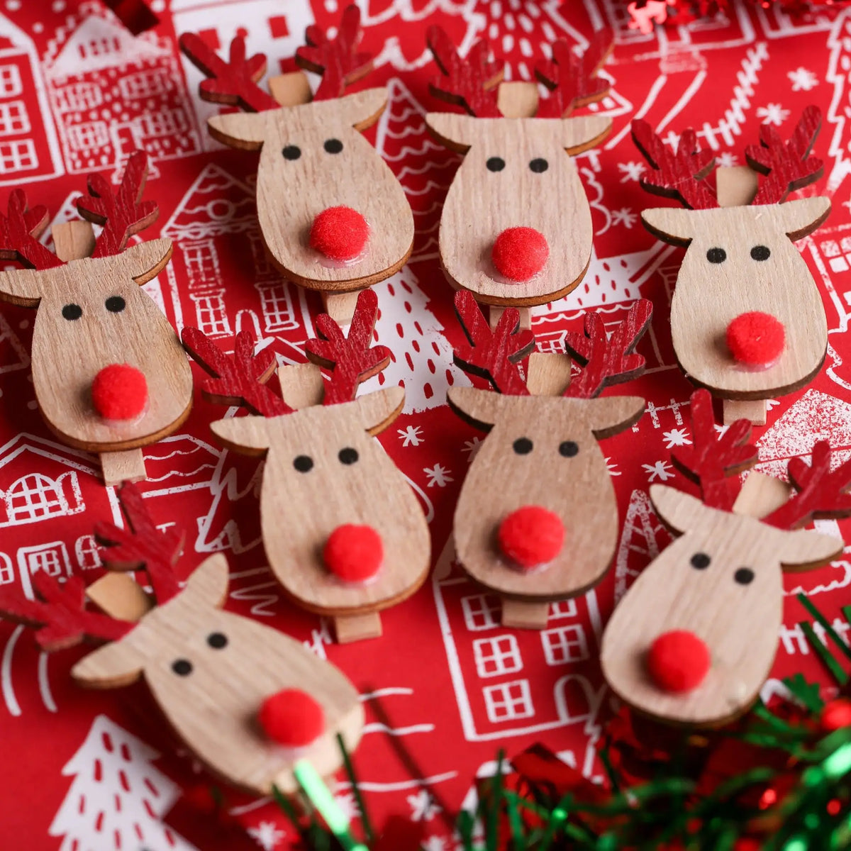 The Printable Heaven Rudolph And Red Nose Clips 8 Pack (XMA4027) features festive wooden reindeer with red noses and antlers, arranged on a red and white background with buildings, trees, and green tinsel—ideal for Christmas crafts.