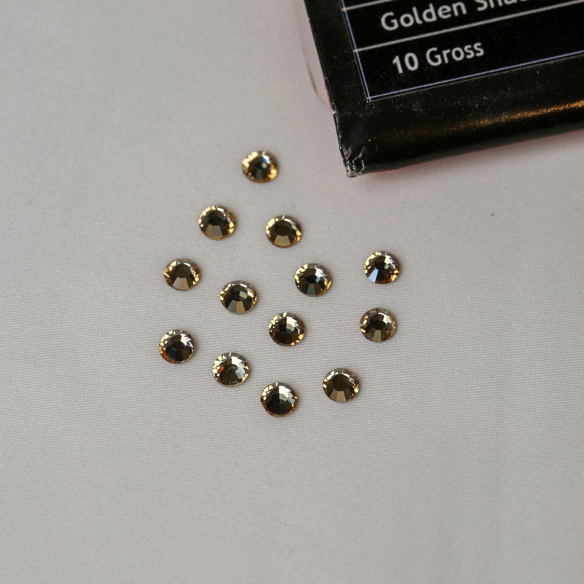 Twelve small, round, golden-brown SS16 Non-Hot Fix Silver Foiled Crystals in Golden Shadow arranged in a cluster next to a black product package.