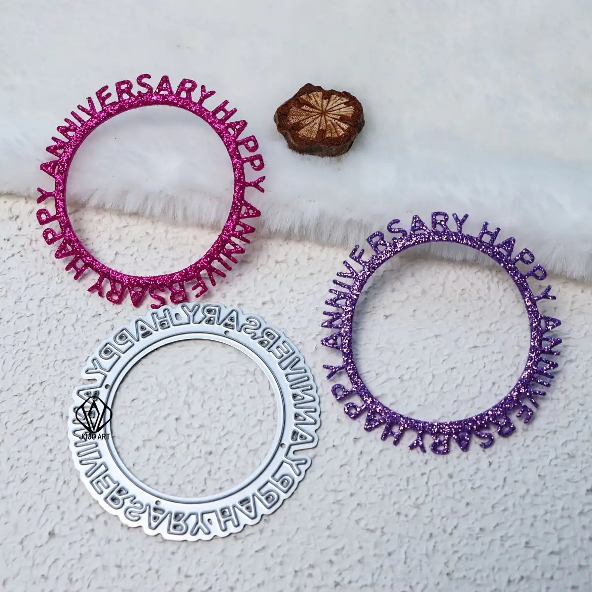 Three round Happy Anniversary cake toppers in pink, purple, and silver glitter are displayed on a white textured surface with a small brown decorative piece nearby—perfect accents for anniversary cards or cardmaking projects using your favorite Printable Heaven Happy Anniversary Cutting Die (1 PC) (W151).