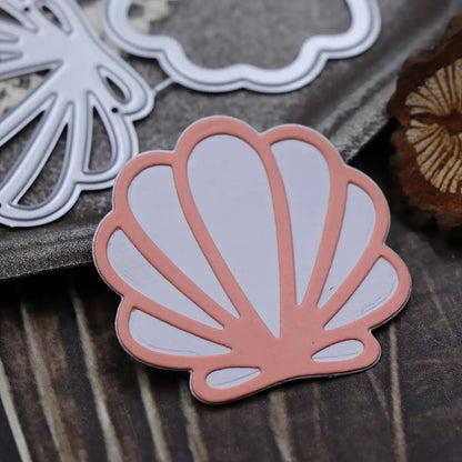 A pink and white seashell-shaped paper cutout rests on a wooden surface next to the Printable Heaven Scallop Seashell Cutting Die (2 PCS) (T234)—an ideal duo for card making or beach-themed crafts. Part of a brown object is visible nearby.