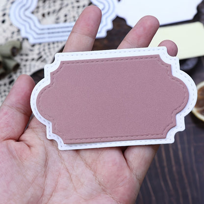 A hand holds a blank, decorative, pink and white label with stitched edges, crafted using the Stitched Edge Nesting Frame Dies – Set of 4 (D124) from Printable Heaven—perfect for card making. Blurred crafting materials can be seen in the background.
