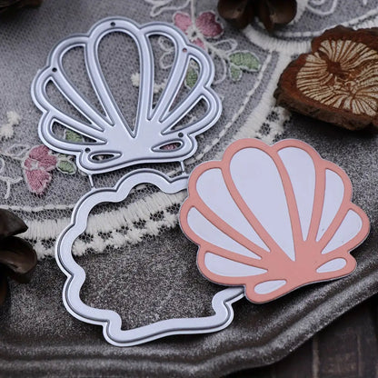 Two Printable Heaven Scallop Seashell Cutting Dies (T234) are displayed on a gray textured surface alongside a pink-and-white paper seashell, with embroidered fabric and wooden accents in the background—ideal for card making and beach-themed crafts.