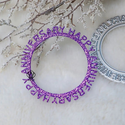 A purple glittery circular piece, created using the Printable Heaven Happy Anniversary Cutting Die (1 PC) (W151), displays the words “HAPPY ANNIVERSARY” and is shown resting on a white furry surface, paired elegantly with frosted branches and part of a silver ornament nearby.