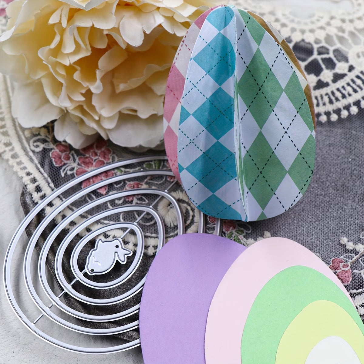 Vibrant Easter paper crafts, featuring paper egg designs created with the Printable Heaven Nesting Eggs with Baby Rabbit Cutting Die (6 PCS) (A097), and a large cream flower are displayed on a lace-trimmed surface adorned with floral patterns.