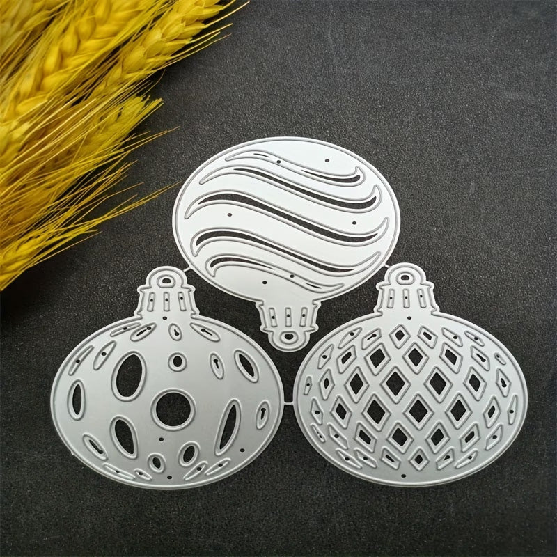 Three Filigree Baubles Cutting Die – Set of 3 (SP020) by Printable Heaven, crafted from silver metal and featuring intricate cutout patterns, are displayed on a dark surface beside a bundle of yellow dried wheat—highlighting the exquisite detail of these dies, which are perfect for bringing elegance to your Christmas craft projects.