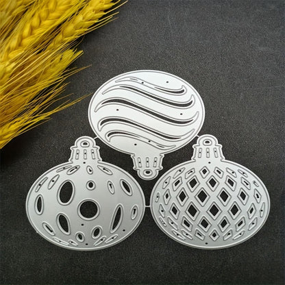 Three Filigree Baubles Cutting Die – Set of 3 (SP020) by Printable Heaven, crafted from silver metal and featuring intricate cutout patterns, are displayed on a dark surface beside a bundle of yellow dried wheat—highlighting the exquisite detail of these dies, which are perfect for bringing elegance to your Christmas craft projects.