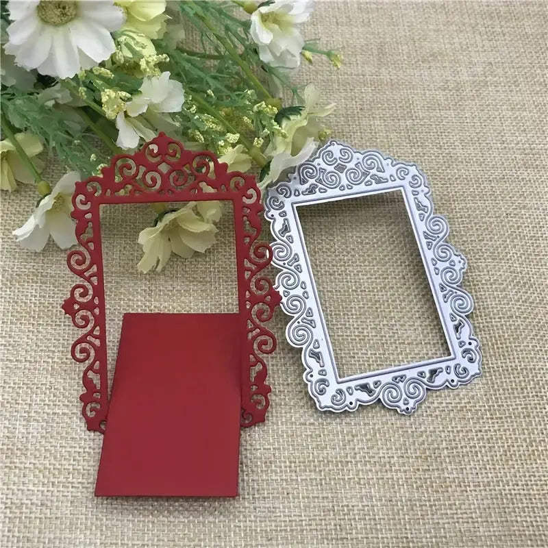 Two ornate rectangular frames, one red and one silver, showcasing intricate scrollwork designs crafted with the Printable Heaven Ornate Decorative Frame Cutting Die (1 PC) (T048), are arranged on a burlap surface beside a bouquet of white and yellow flowers. Perfect as cardmaking embellishments, these elegant frames bring an added touch of sophistication to any creative project.