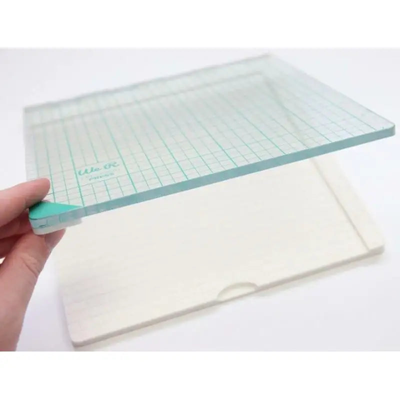 A hand holds a transparent glass board with a teal grid and rounded corners over a rectangular white base, both items set against a white background—an ideal arrangement for using the Printable Heaven We R Memory Keepers WR Tool Precision Press (WR663007).