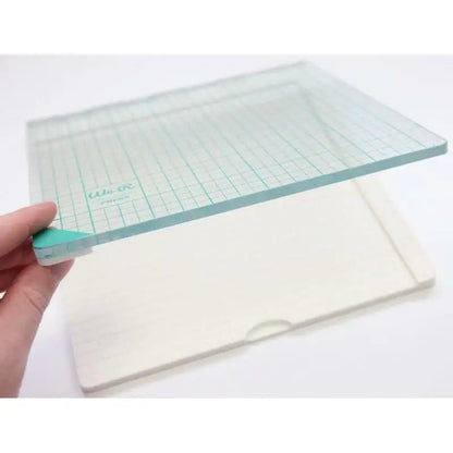 A hand holds a transparent glass board with a teal grid and rounded corners over a rectangular white base, both items set against a white background—an ideal arrangement for using the Printable Heaven We R Memory Keepers WR Tool Precision Press (WR663007).