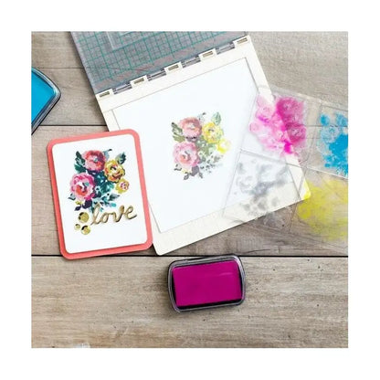 Handmade card featuring painted flowers and a love sentiment displayed alongside crafting supplies, ink pads, a Printable Heaven We R Memory Keepers WR Tool Precision Press (WR663007), and a floral stencil on a wooden surface.